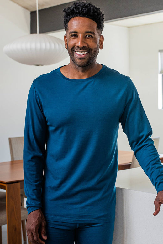 A man standing and smiling, leaning against a counter wearing Yala Superfine Merino Wool Long Sleeve Top in Lapis