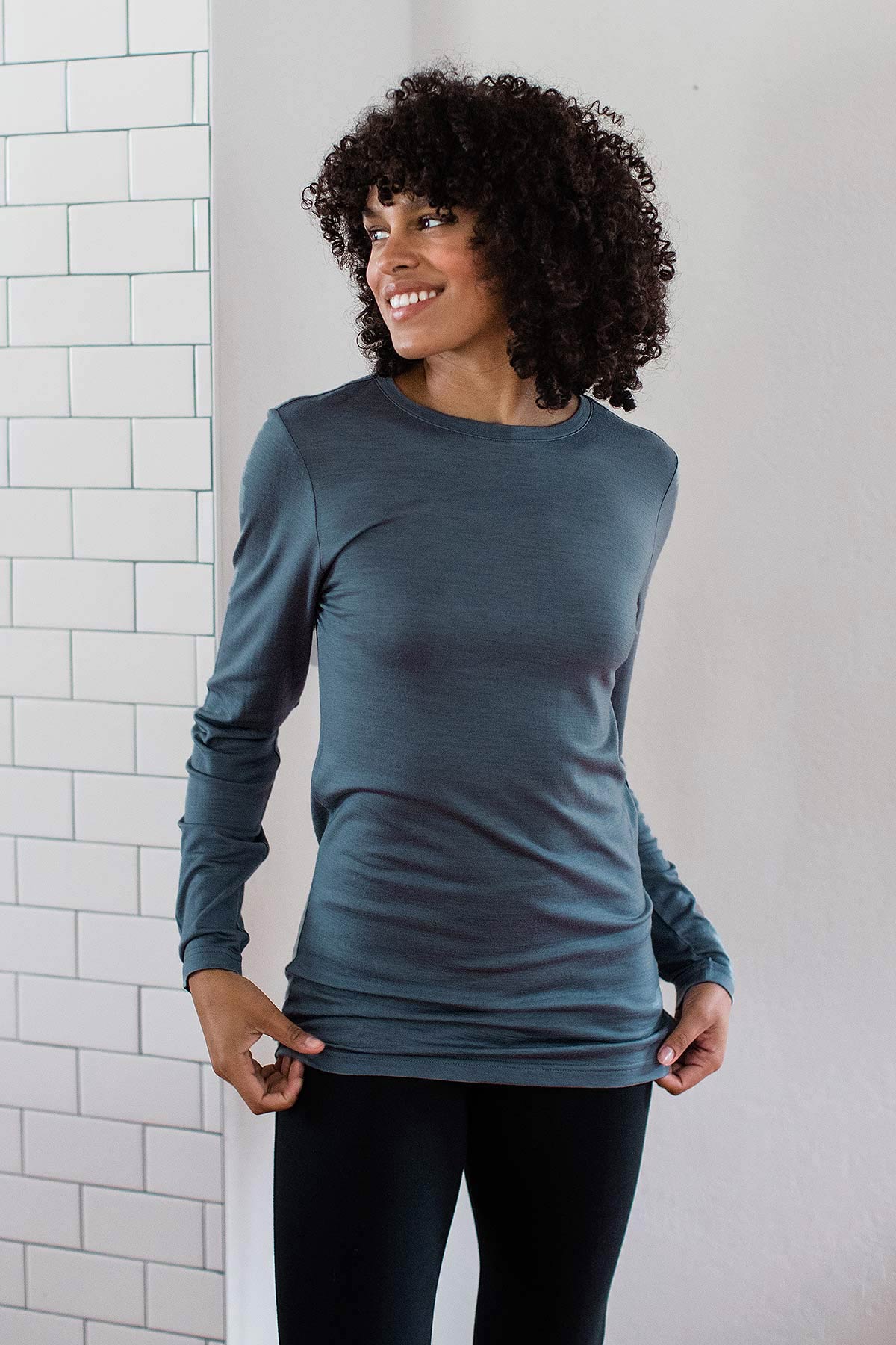 A woman standing and smiling while adjusting the edge of her top, wearing Yala Superfine Merino Wool Long Sleeve Top in Storm Grey