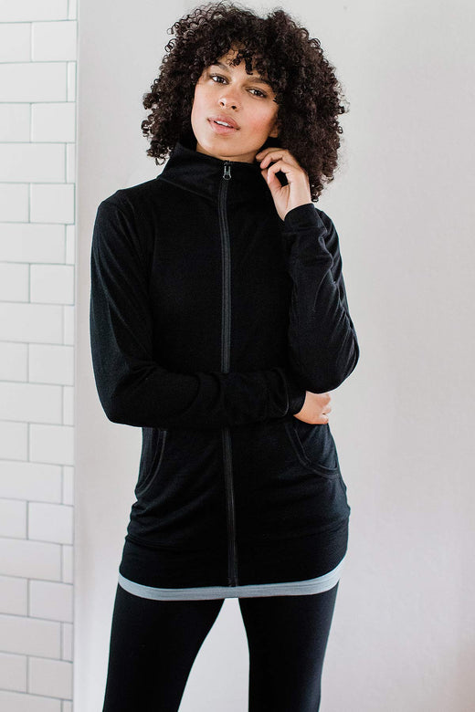 A womand standing with one hand holding her waist and the other raised to adjust her jacket collar, wearing Yala Superfine Merino Wool Track Jacket in Black