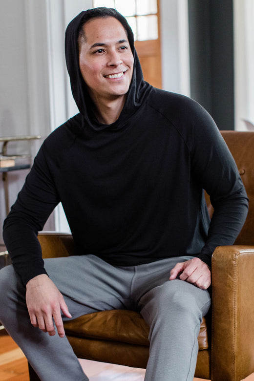 A amn sitting in a chair and smiling while looking off to the side, wearing Yala Travis Men's Hooded Long Sleeve Raglan Bamboo Tee Shirt in Black