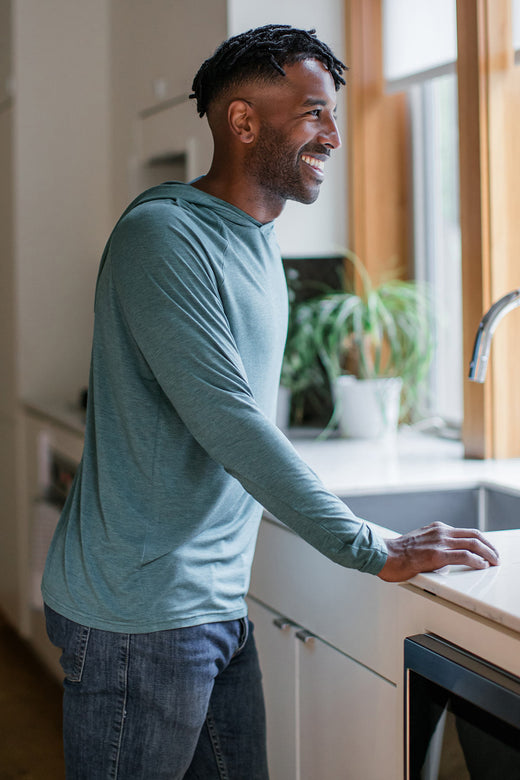 A man standing and smiling while leaning forward with his hands against a countertop, wearing Yala Travis Hooded Long Sleeve Raglan Bamboo Tee shirt in Teal Melange