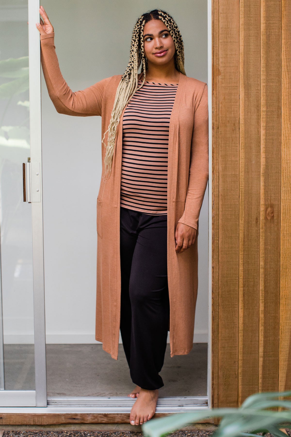 A woman standing in a sliding glass doorway, one hand resting on the doorframe, wearing Yala Brooke Cardigan Duster Bamboo Sweater in Camel