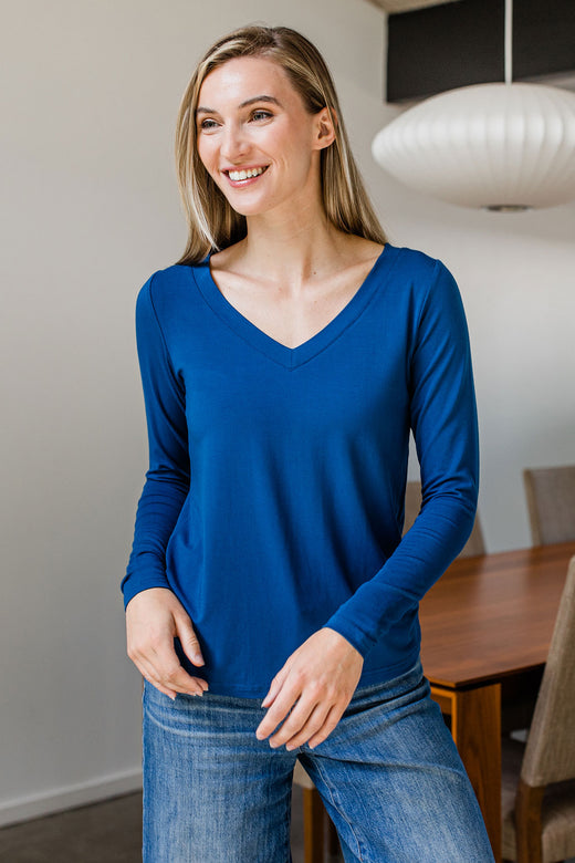A woman standing and smiling with her hands in front of her waist, wearing Yala Jill V-Neck Long Sleeved Bamboo Top in Lapis