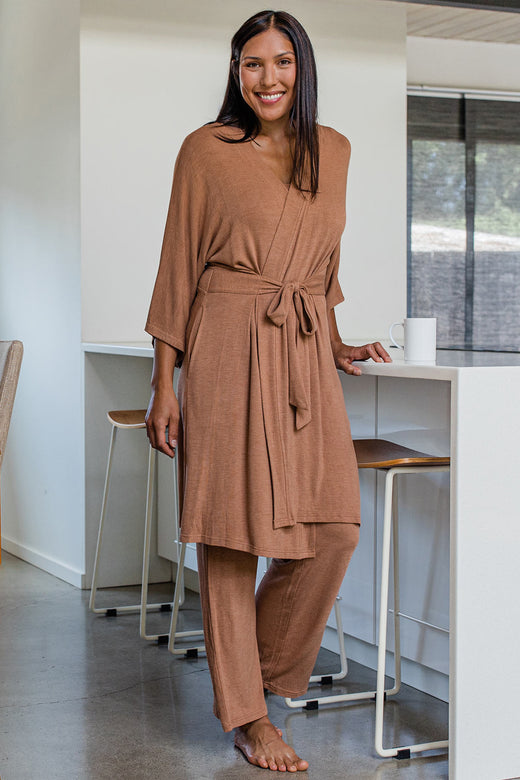 A womand standing and smiling with one hand on a countertop, wearing Yala Kendra Kimono Bamboo Sweater Robe in Camel