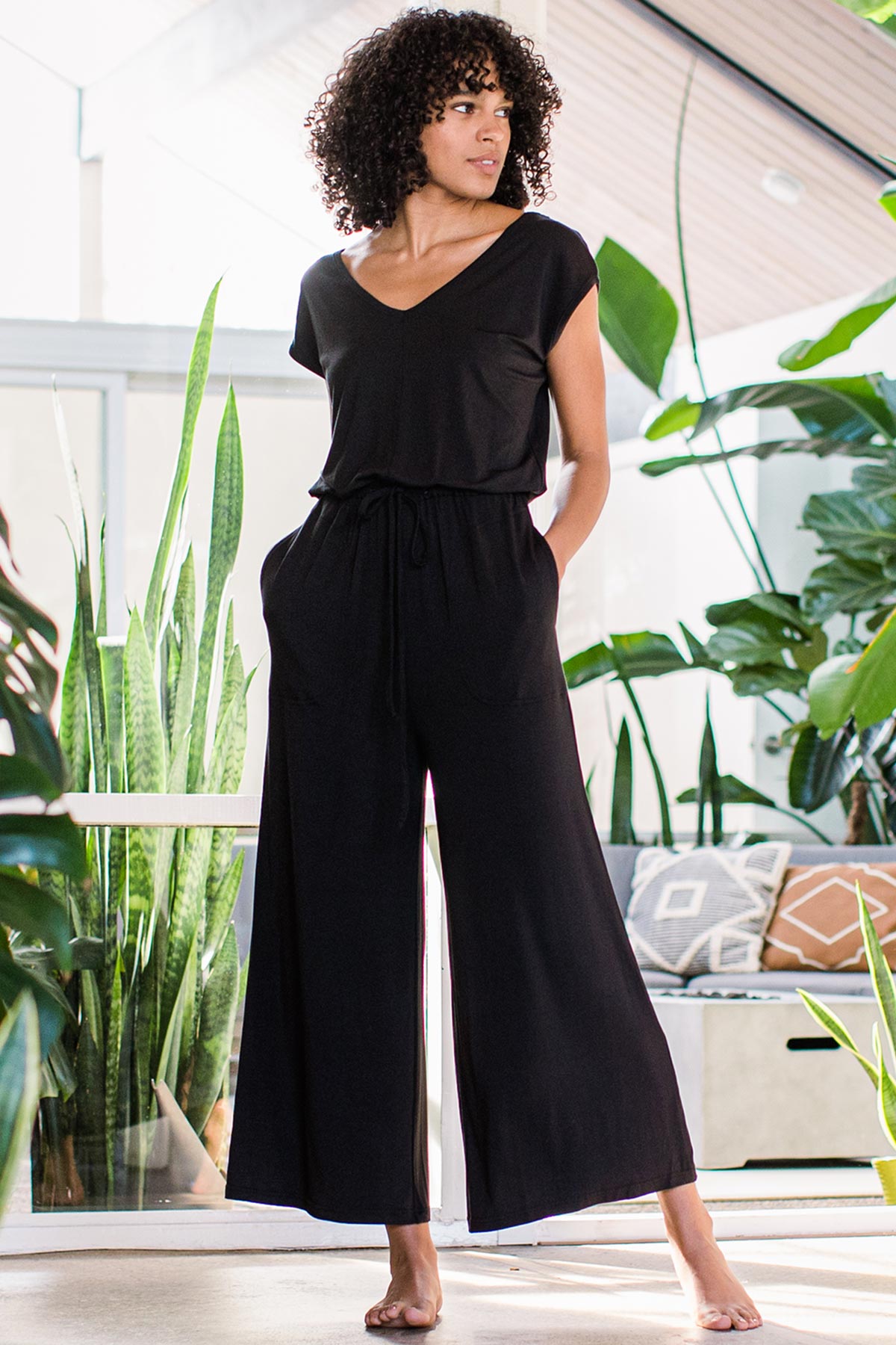 A woman standing and gazing to the side with both hands in her pockets, wearing Yala Kiova V-Neck Bamboo Jumpsuit in Black