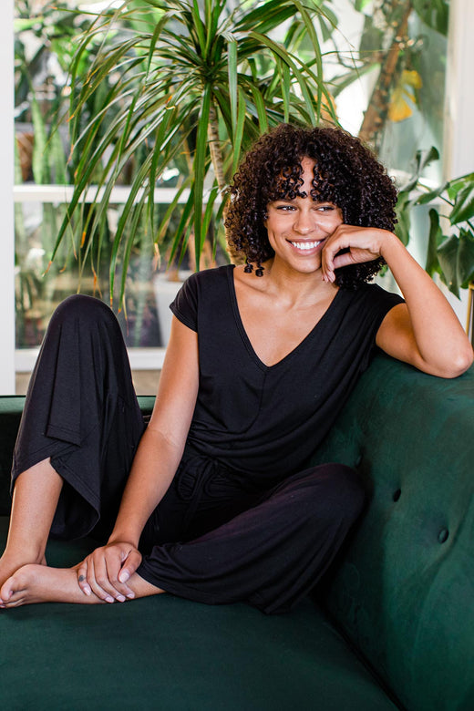 A woman sitting on a couch and smiling, wearing Yala Kiova V-Neck Bamboo Jumpsuit in Black