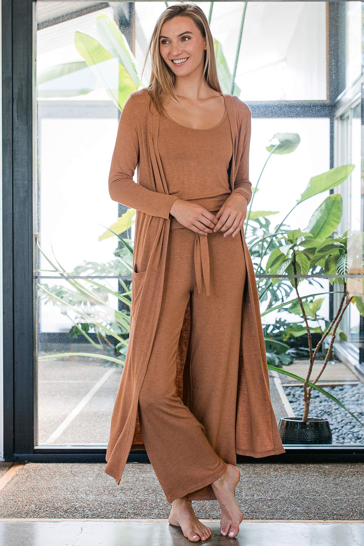 A woman standing and smiling with one leg crossed in front, wearing Yala Peyton Bamboo Sweater Drawstring Lounge Pants in Camel