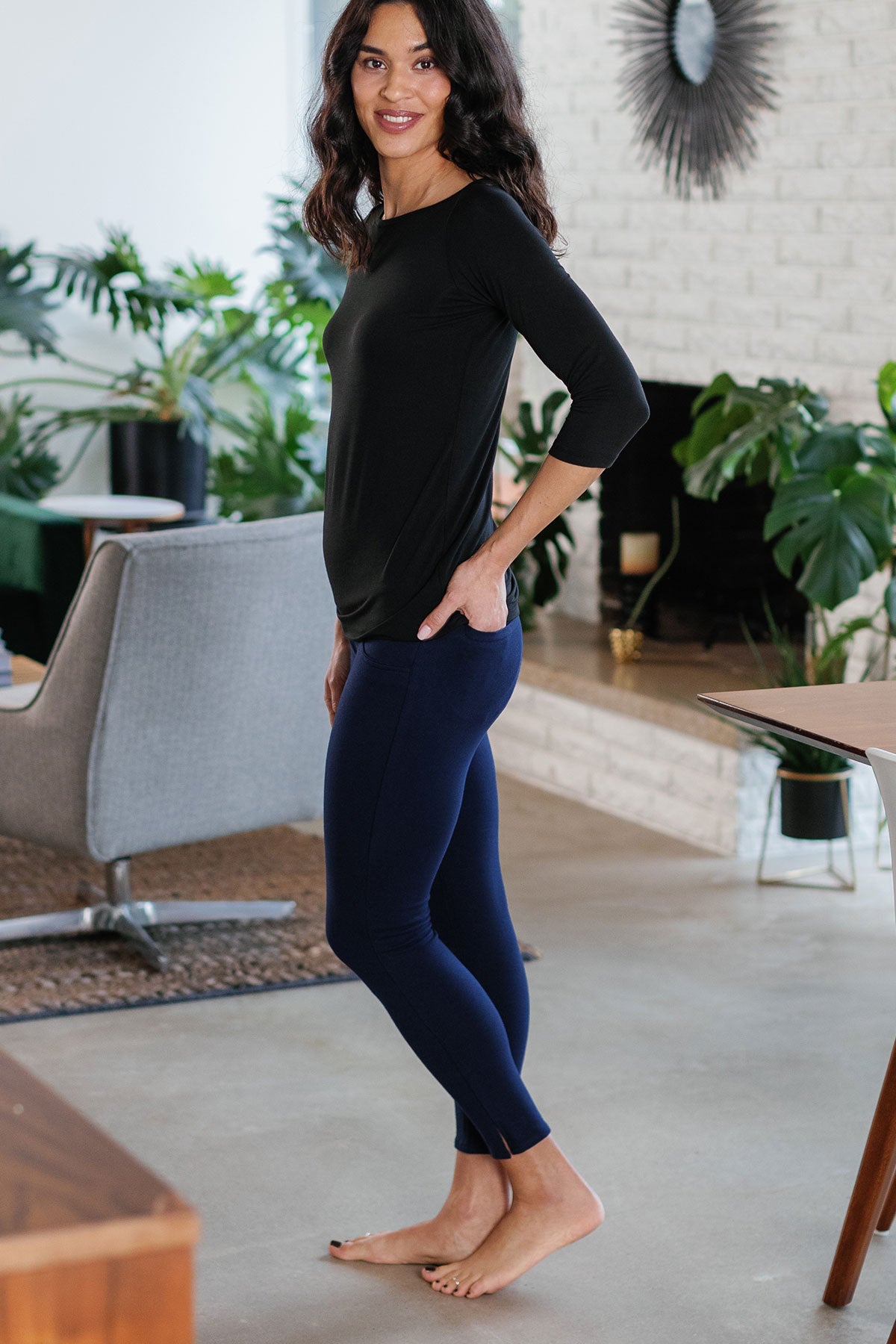 A woman standing facing to the side, looking towards the camera, wearing Yala Abby Mid-Waist Cropped Bamboo and Organic Cotton Pencil Pants in Navy