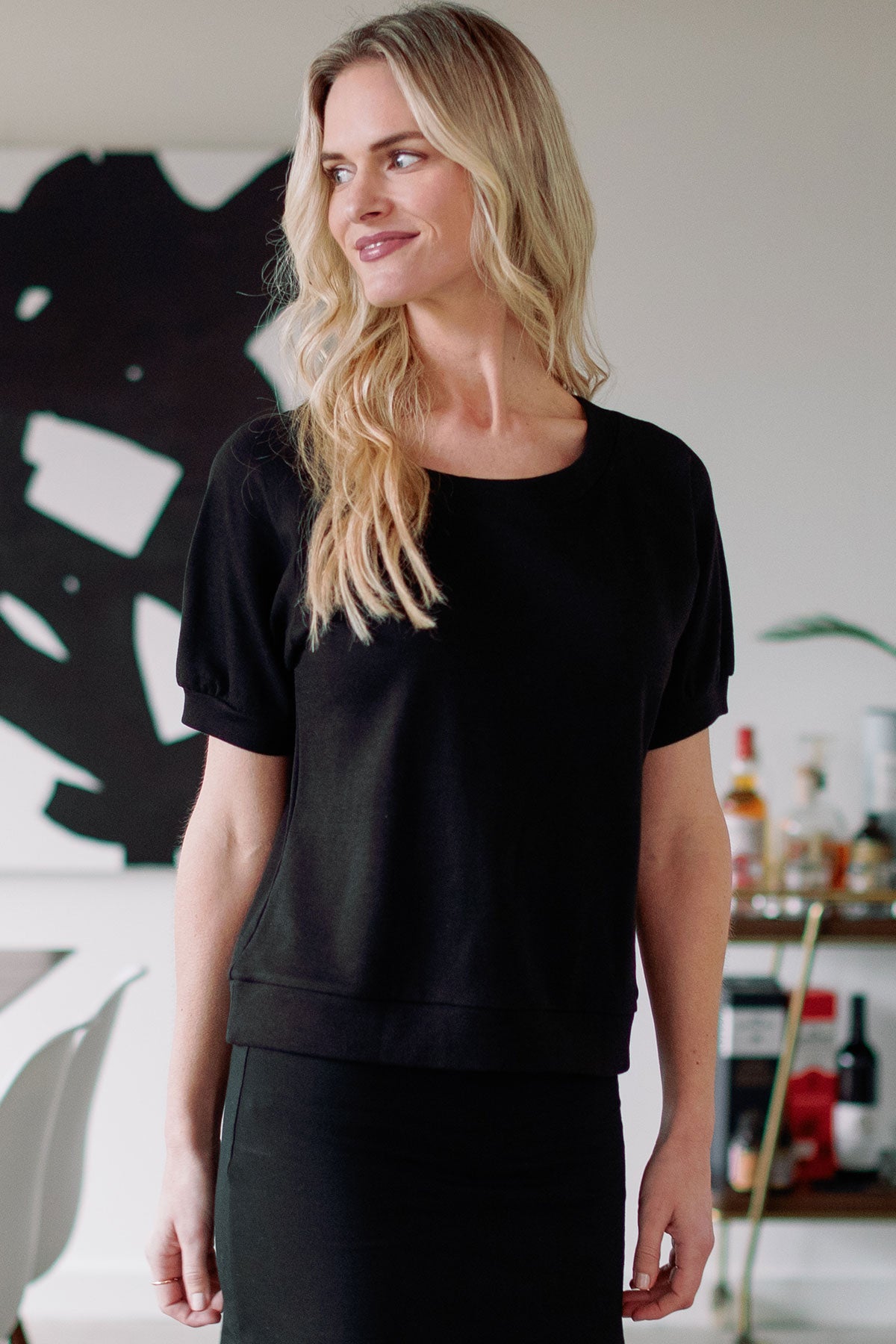 A woman standing and smiling while looking to the side, wearing Yala Berkeley Puff Short Sleeve Bamboo and Organic Cotton Raglan Sweatshirt Top in Black