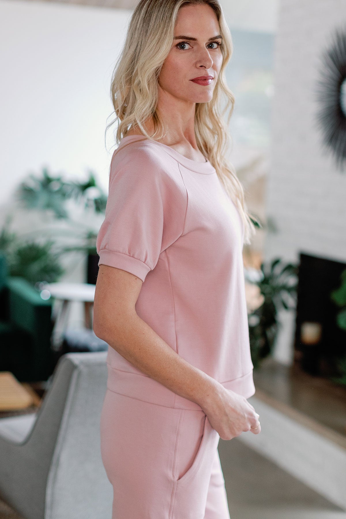 A woman standing facing to the side, looking towards the camera with hands at her waistline, wearing Yala Berkeley Puff Short Sleeve Bamboo and Organic Cotton Raglan Sweatshirt Top in Lotus Pink