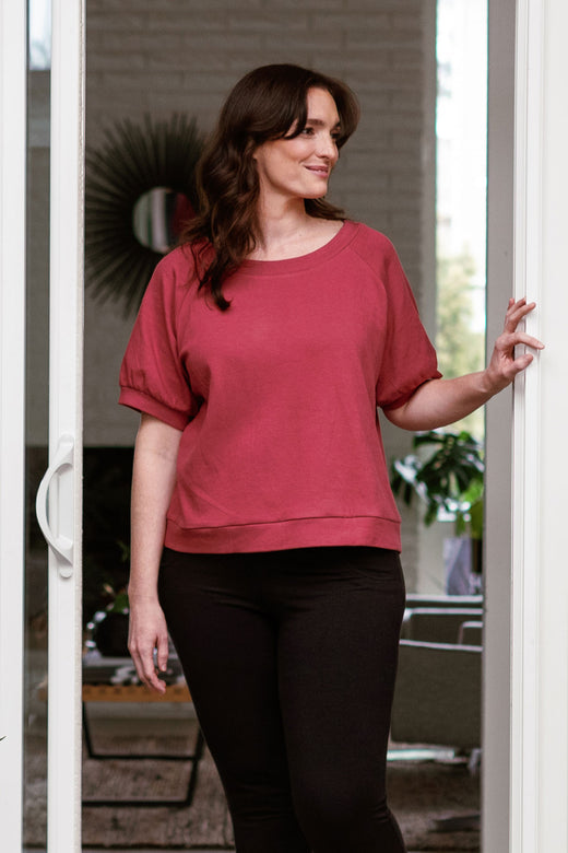 A woman standing in a sliding glass doorway, smiling and looking to the side, wearing Yala Berkeley Puff Short Sleeve Bamboo and Organic Cotton Raglan Sweatshirt Top in Rosewood