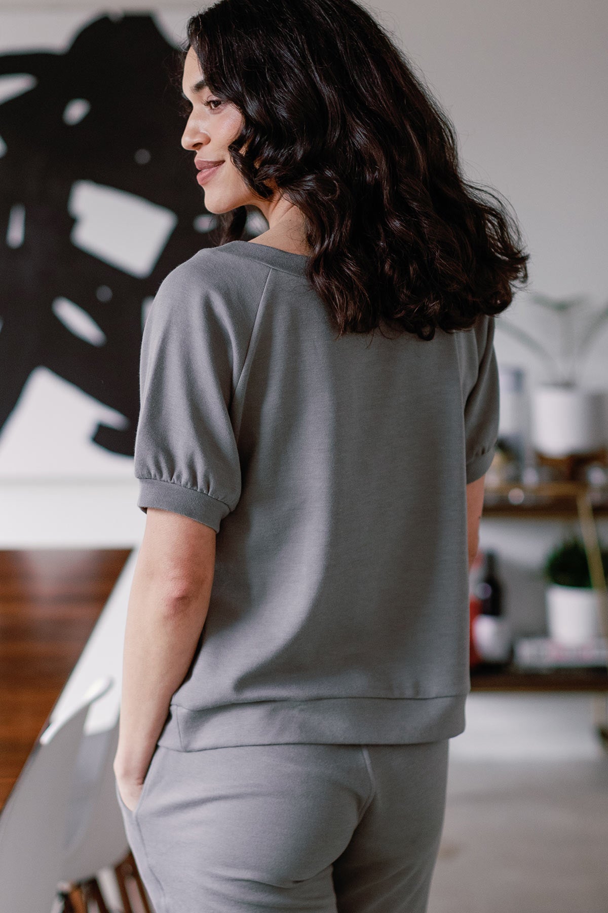 A woman standing facing away from the camera, looking to the side, wearing Yala Berkeley Puff Short Sleeve Bamboo and Organic Cotton Raglan Sweatshirt Top in Space Grey