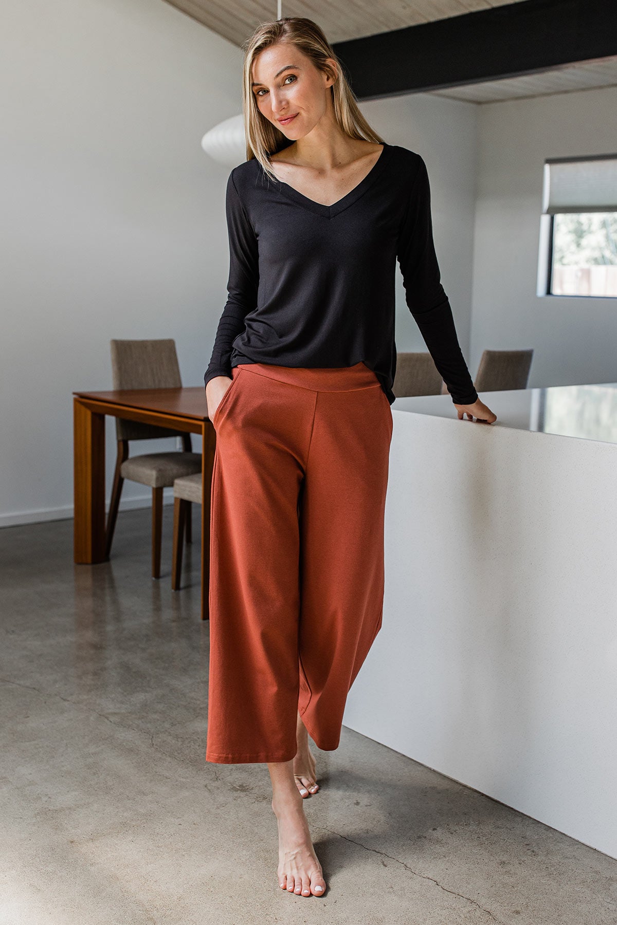 A woman stands leaning on a counter with a hand in her pocket, wearing Yala Billie Ultra-Stretch Bamboo and Organic Cotton Classic Wide Leg Cropped Pants
