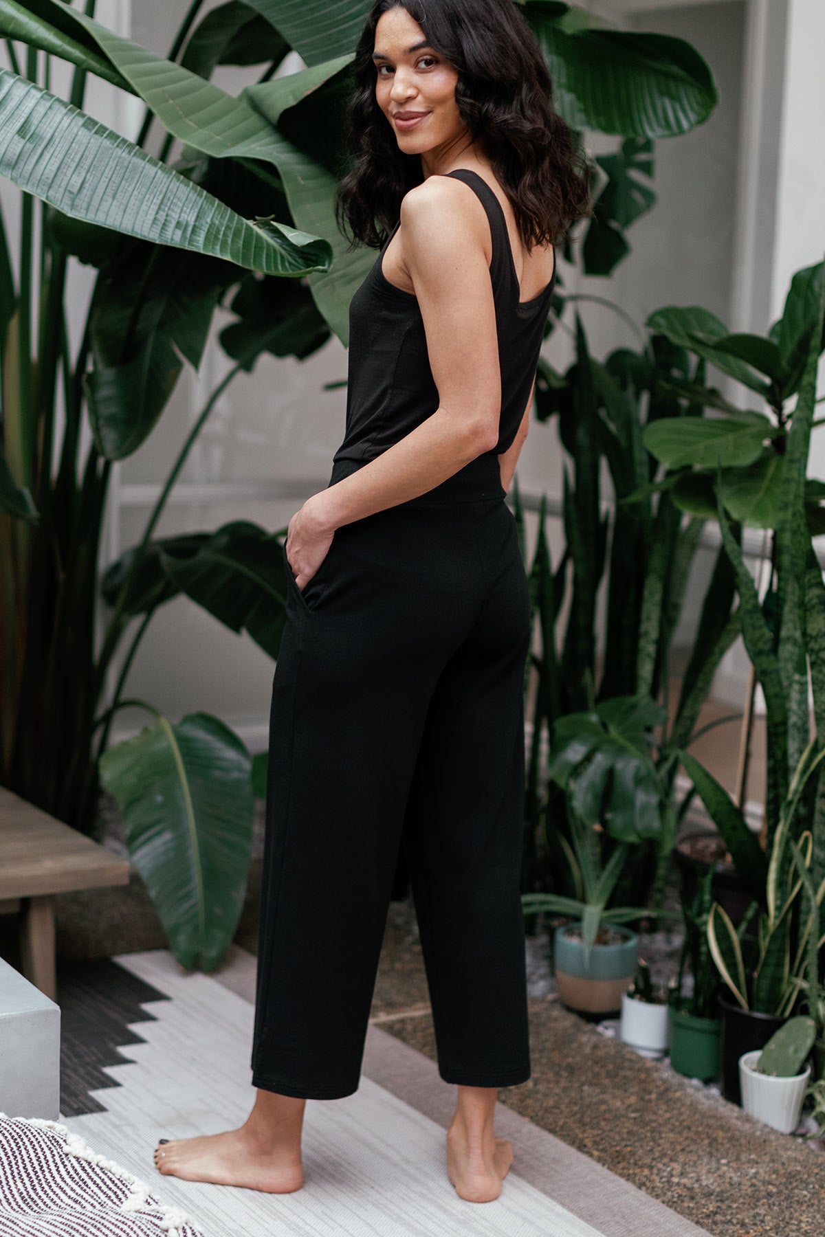 A woman standing facing away and turning her waist to smile at the camera, wearing Yala Billie Ultra-Stretch Bamboo and Organic Cotton Classic Wide Leg Cropped Pants in Black