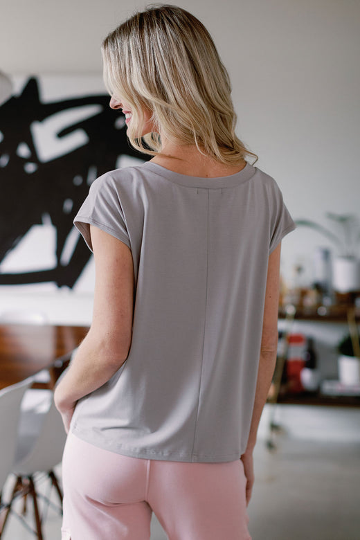 A woman standing facing away from the camera, smiling to the side, wearing Yala Dakota V-Neck Cap Sleeve Bamboo Top in Ash
