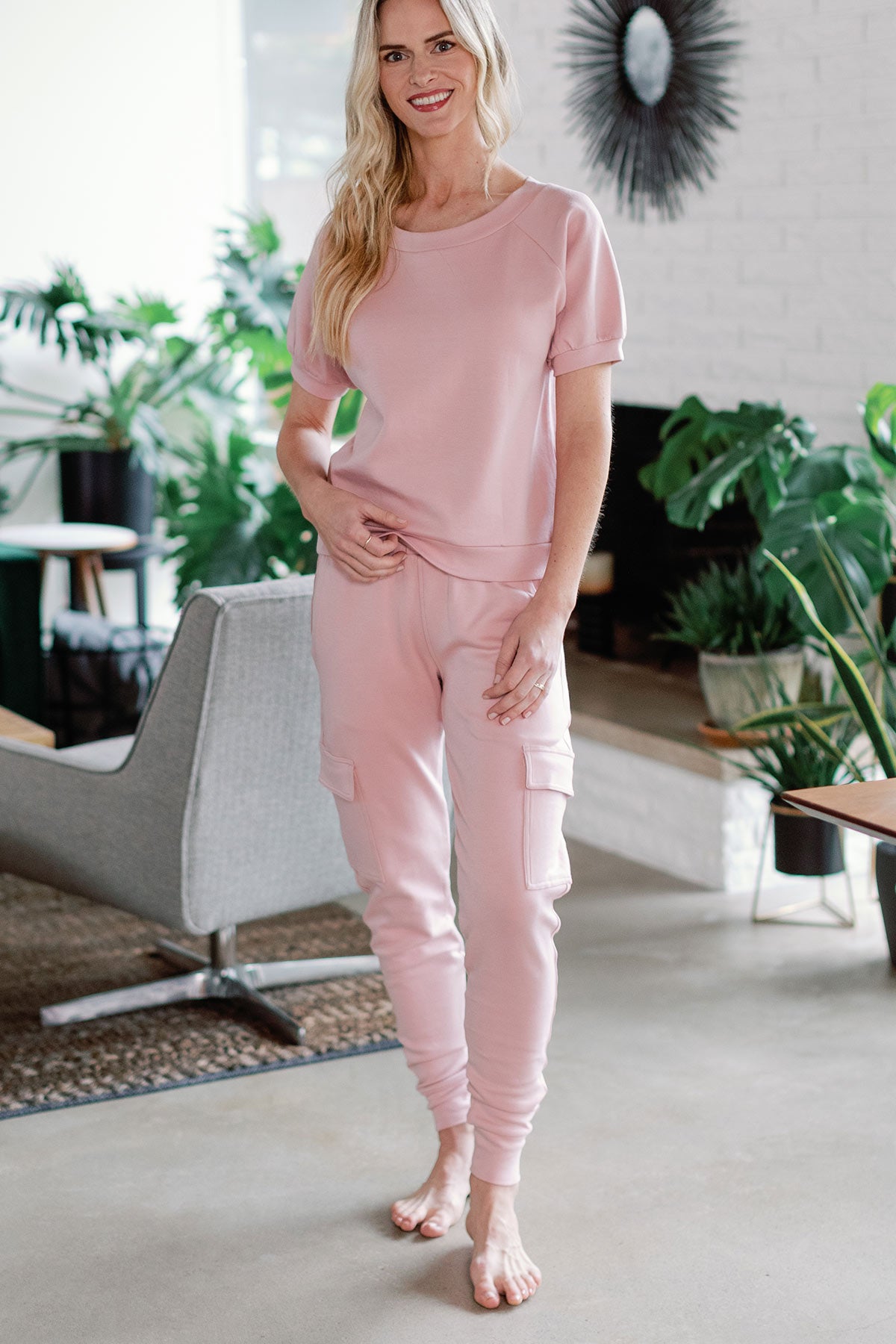 A woman standing and smiling with one hand resting at her beltline, wearing Yala Fay Bamboo and Organic Cotton Cargo Joggers in Lotus Pink