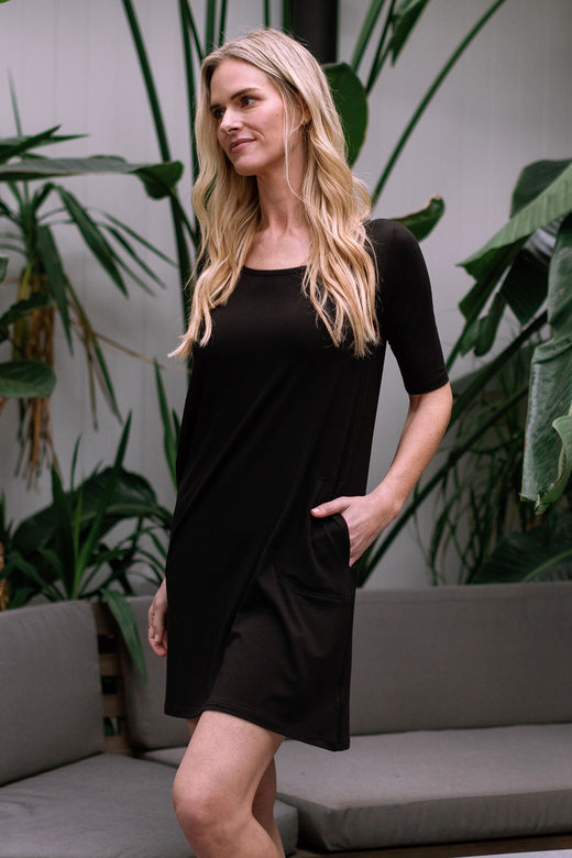 A woman standing facing partly to the side with one hand in her pocket, wearing Yala Frida Relaxed Fit Scoop Neck Short Sleeve Bamboo Lounge Dress in Black