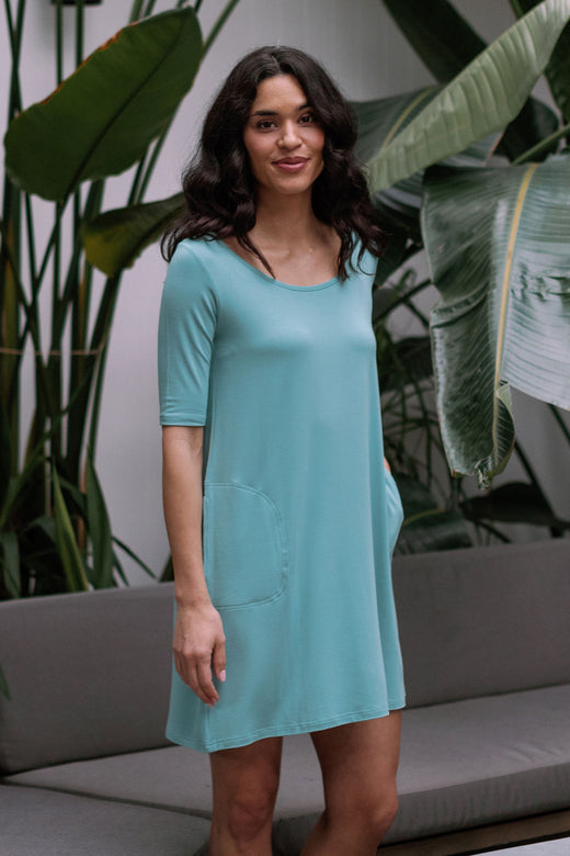 A woman standing with one hand in her pocket, wearing Yala Frida Relaxed Fit Scoop Neck Short Sleeve Bamboo Lounge Dress in Nile