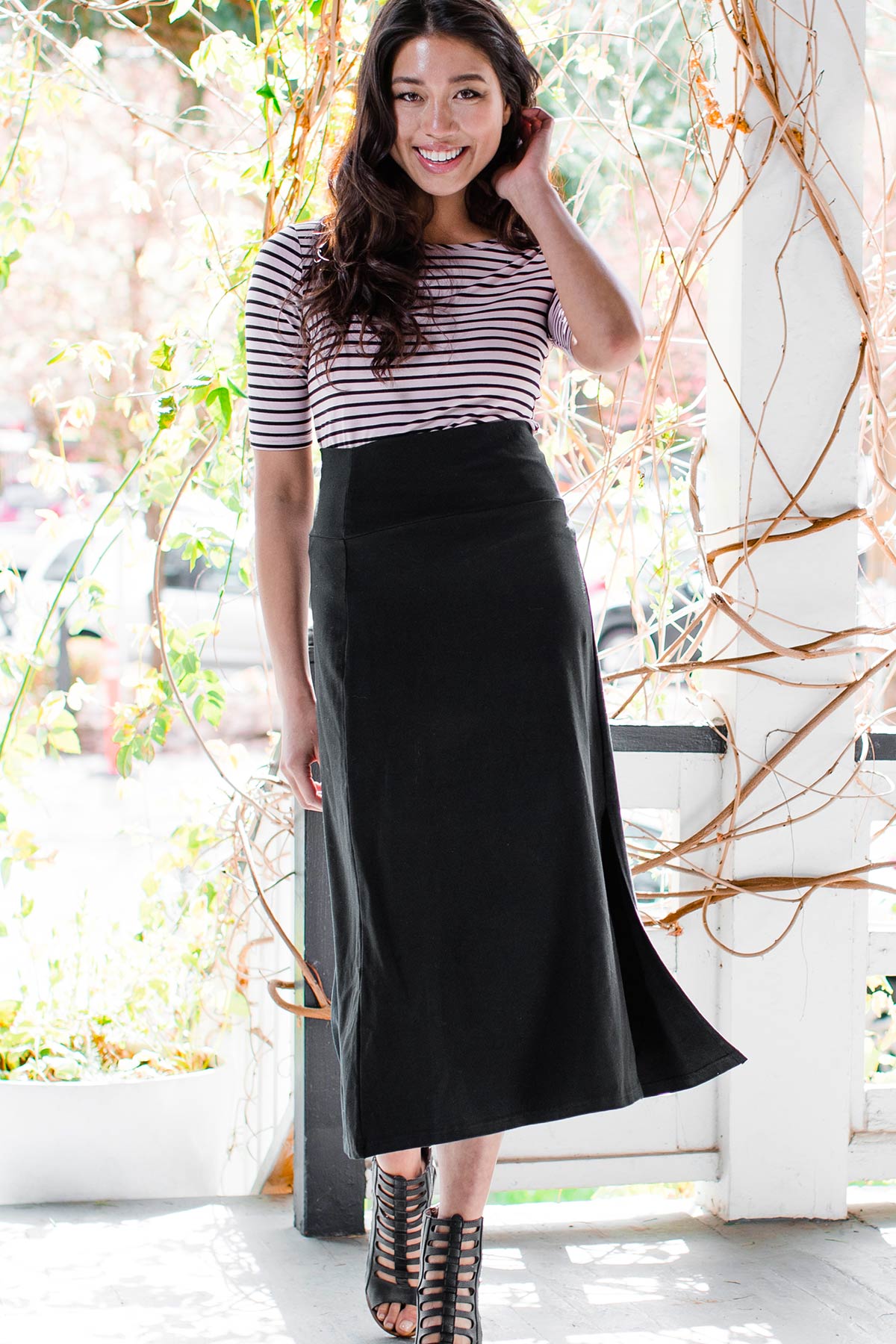 A woman walking forward and smiling while brushing her hair aside with one hand, wearing Yala Hannah Ultra-Stretch High-Waisted Bamboo and Organic Cotton Midi Skirt in Black