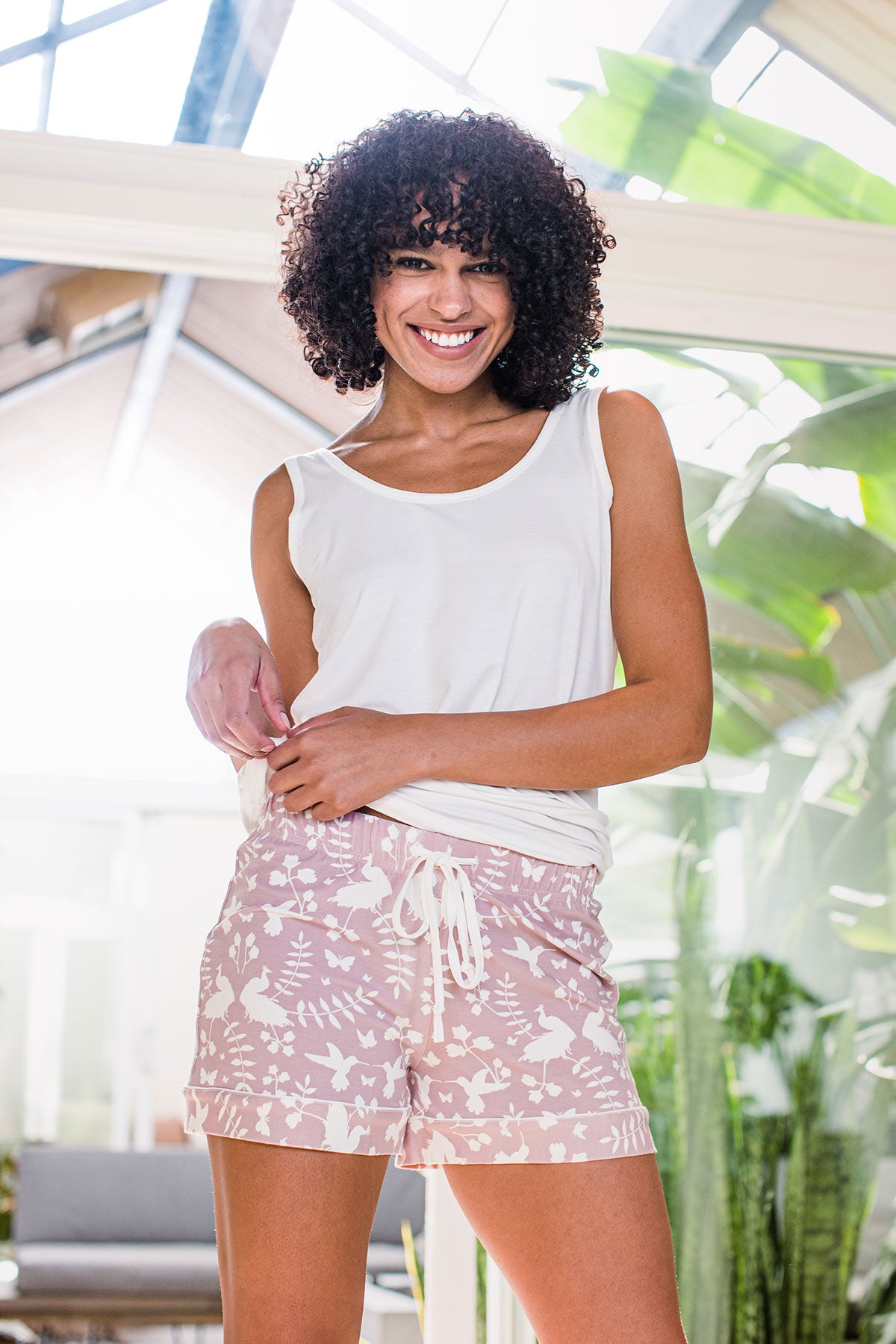 A woman standing and smiling with both hands holding the hem of her top, wearing Yala Lauren Piped Bamboo Pajama Shorts in Secret Garden