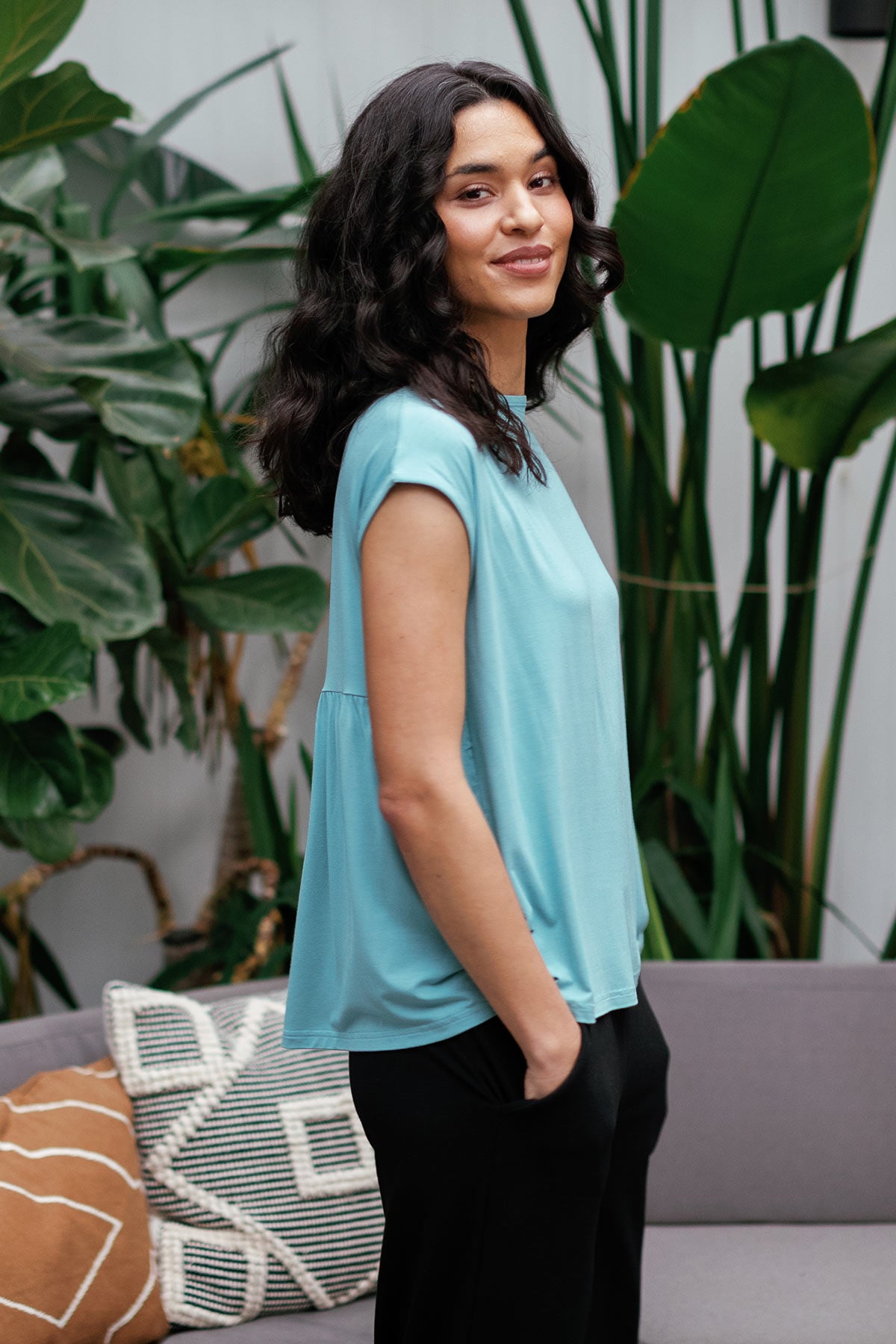 A woman standing facing to the side, hands in pockets and looking towards the camera, wearing Yala Opal Swing Bamboo Top in Nile