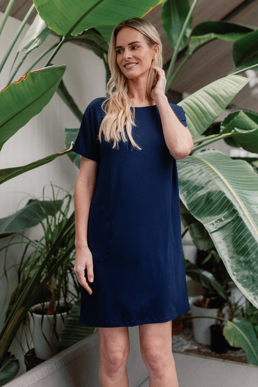 A woman standing and looking to the side with one hand brusing her hair aside, wearing Yala Reese T-Shirt Bamboo Shift Dress in Navy