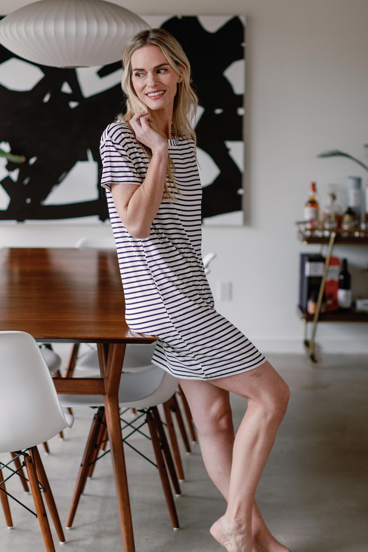 A woman sitting on the edge of a table and smiling over her shoulder, wearing Yala Reese T-Shirt Bamboo Shift Dress in Navy Newport Stripe