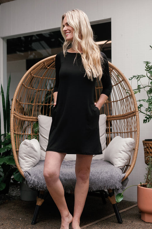 A woman standing and smiling while gazing to the side with on knee bent, wearing Yala Rita Boatneck A-Line Bamboo Dress With Pockets in Black
