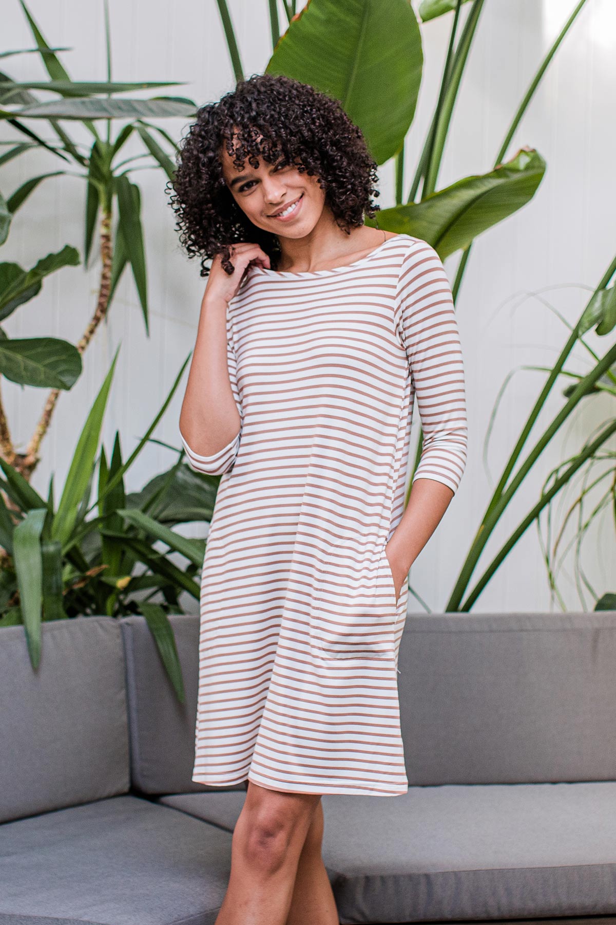 A woman standing and smiling with one leg crossed in front, wearing Yala Rita Boatneck A-Line Bamboo Dress With Pockets in Camel Newport Stripe