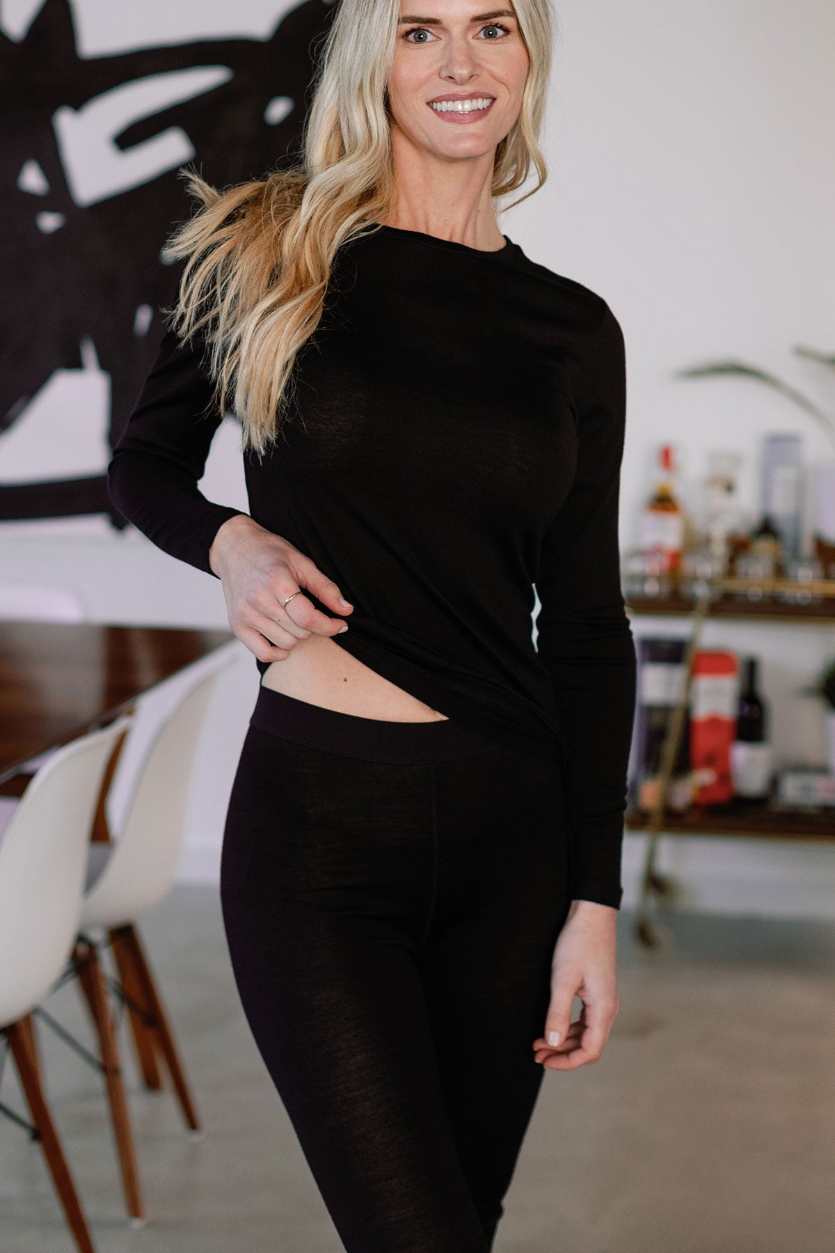 A woman standing and lifting the edge of her top, wearing Yala Superfine Merino Wool Leggings in Black